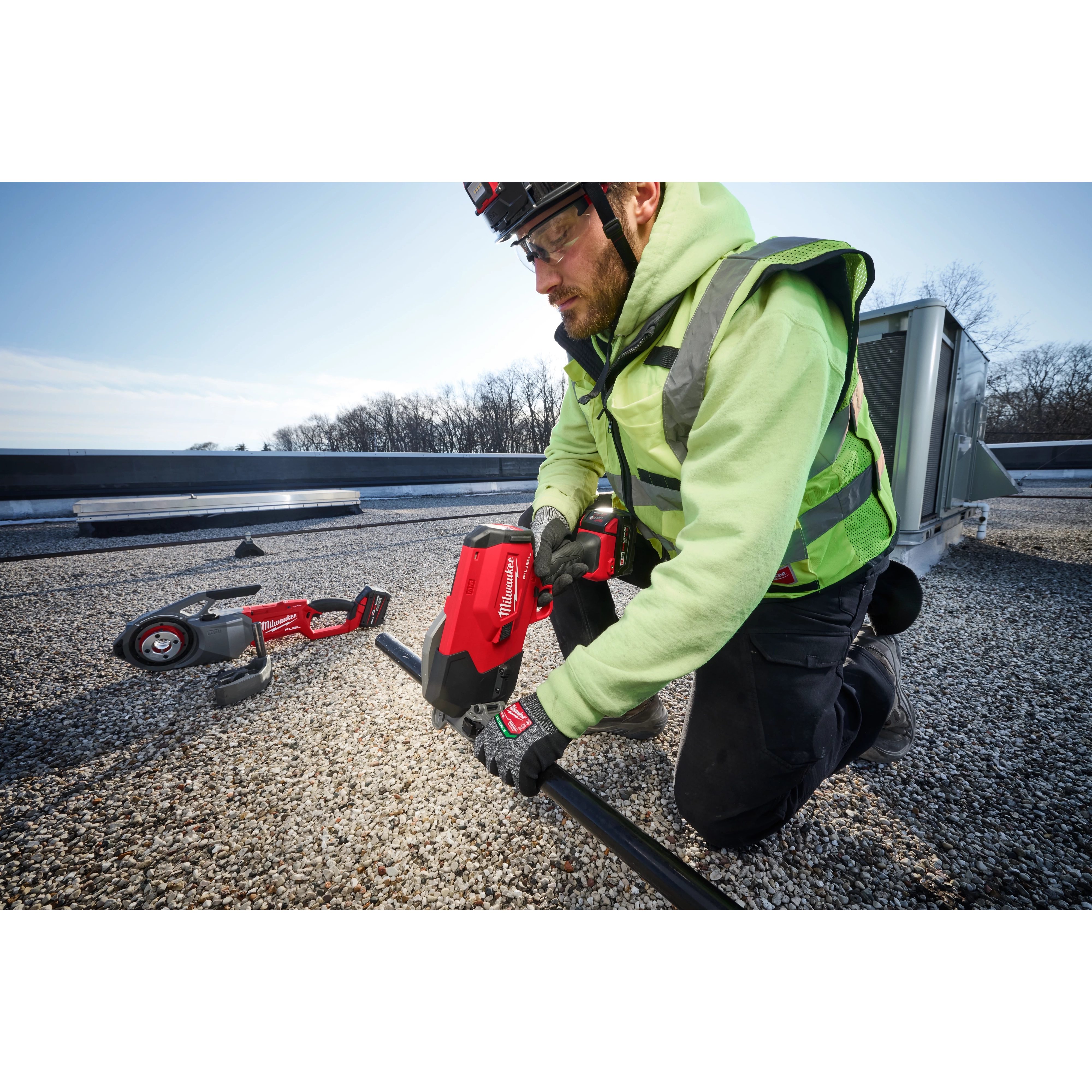 A person in a high-visibility jacket uses an M18 FUEL™ ½”-1” Steel Pipe Cutter to cut through a pipe on a gravel rooftop. Another tool lies on the ground nearby. It's a bright day with a clear sky and some trees in the background.