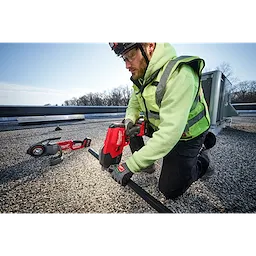 A person in a high-visibility jacket uses an M18 FUEL™ ½”-1” Steel Pipe Cutter to cut through a pipe on a gravel rooftop. Another tool lies on the ground nearby. It's a bright day with a clear sky and some trees in the background.