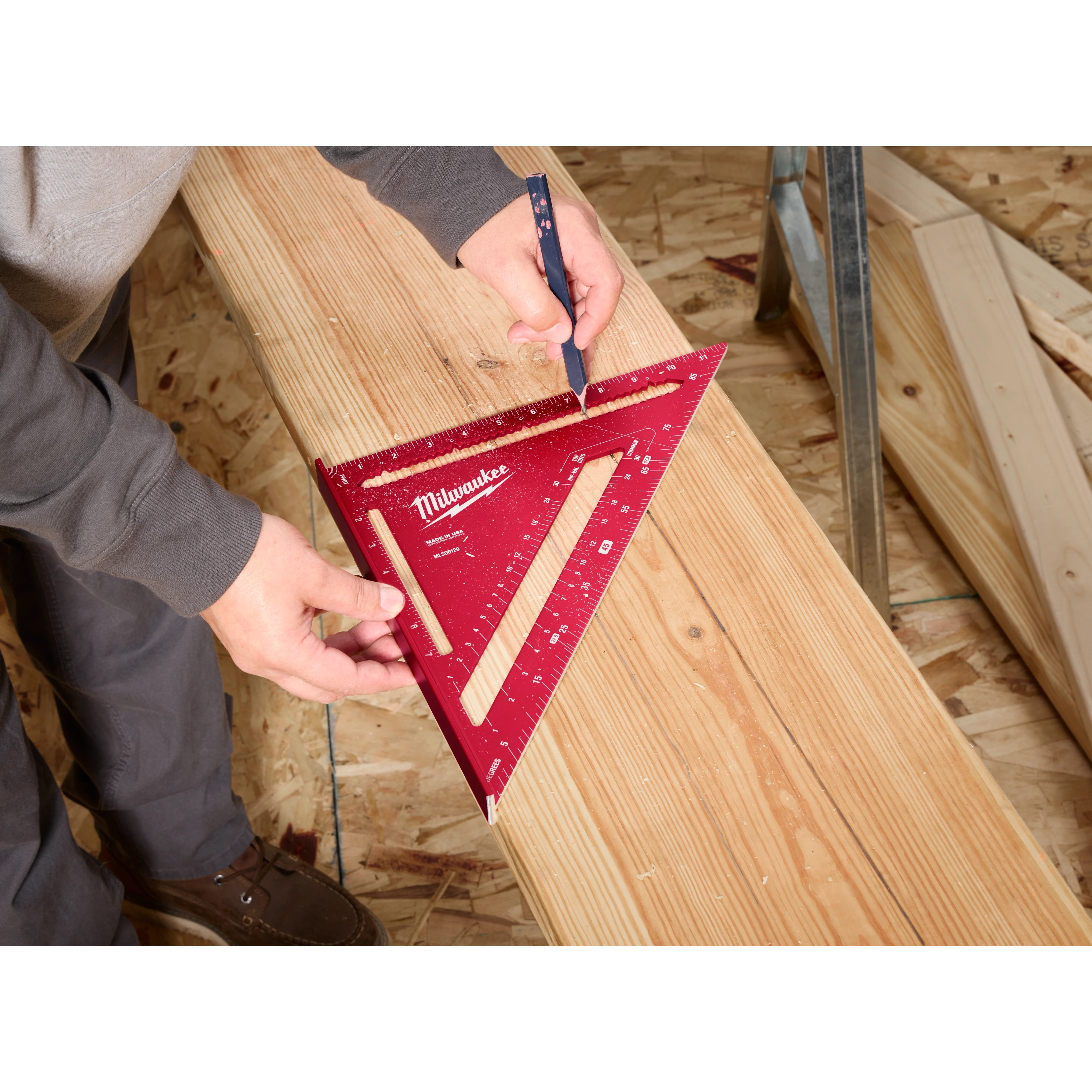 A person using a 12" Rafter Square to draw a line on a wooden board. The 12" Rafter Square is red with measurement markings and the Milwaukee brand name.