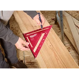 A person using a 12" Rafter Square to draw a line on a wooden board. The 12" Rafter Square is red with measurement markings and the Milwaukee brand name.