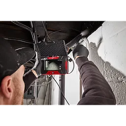 A person is using an M12™ Wireless Monitor to inspect a pipe near a wall. The device has a red and black casing and is displaying the interior view of the pipe on its screen. The individual is wearing gloves and a black cap.