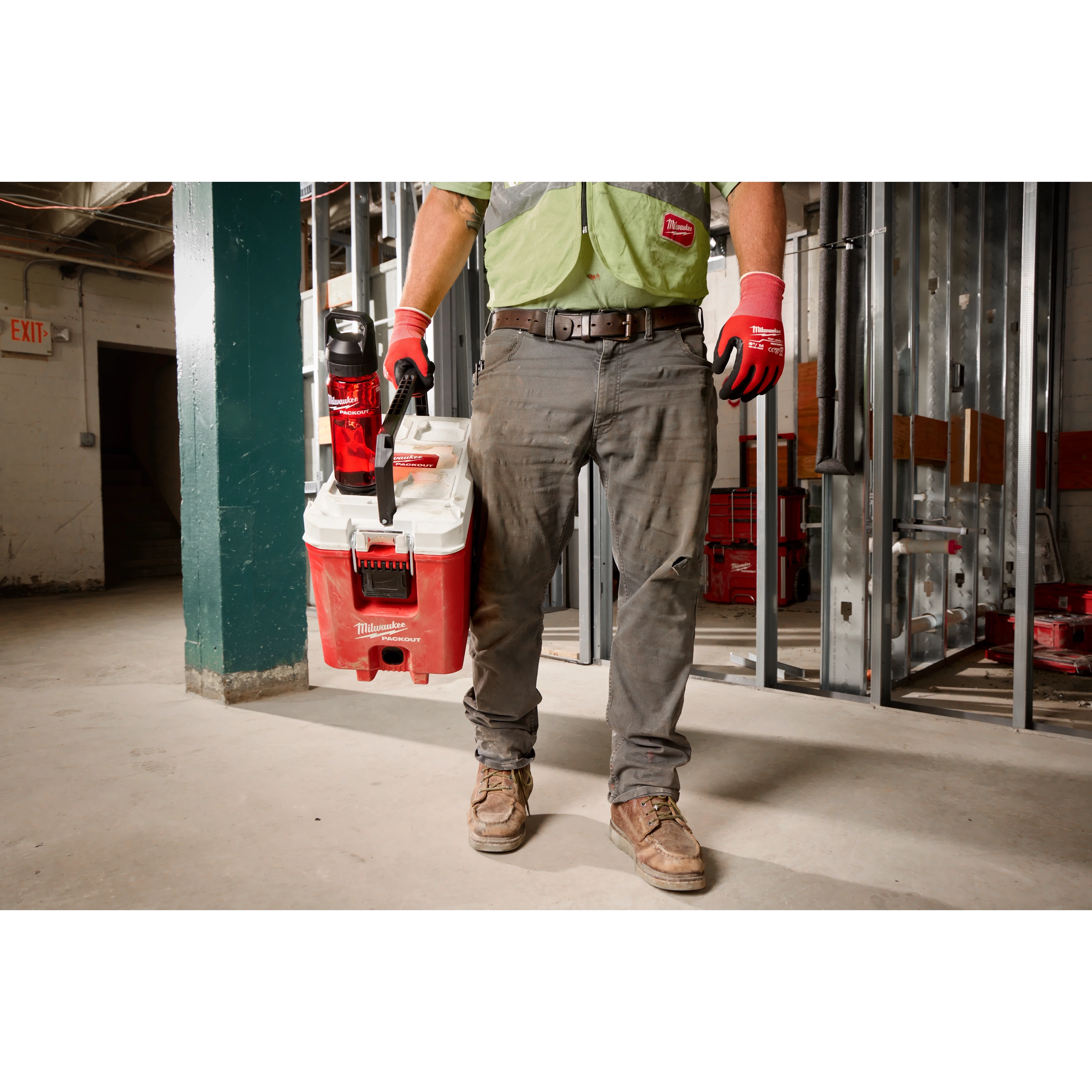 A person wearing work gloves and a green high-visibility vest carries a Milwaukee PACKOUT Cooler in one hand and a PACKOUT 25oz Bottle with Chug Lid in the other