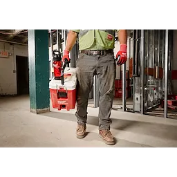 A person wearing work gloves and a green high-visibility vest carries a Milwaukee PACKOUT Cooler in one hand and a PACKOUT 25oz Bottle with Chug Lid in the other