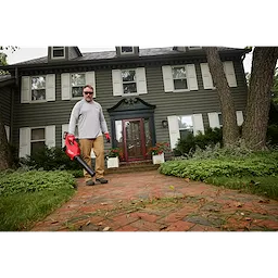 A person stands on a brick walkway in front of a green, two-story house holding an M18™ Brushless Blower. The blower is red with a black nozzle. Green bushes and trees surround the house, and fallen leaves are visible on the walkway.