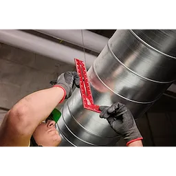 A person wearing gloves is using an 8"x12" Framing Square to measure or mark a large metal cylindrical duct. The framing square is red with white measurement markings.