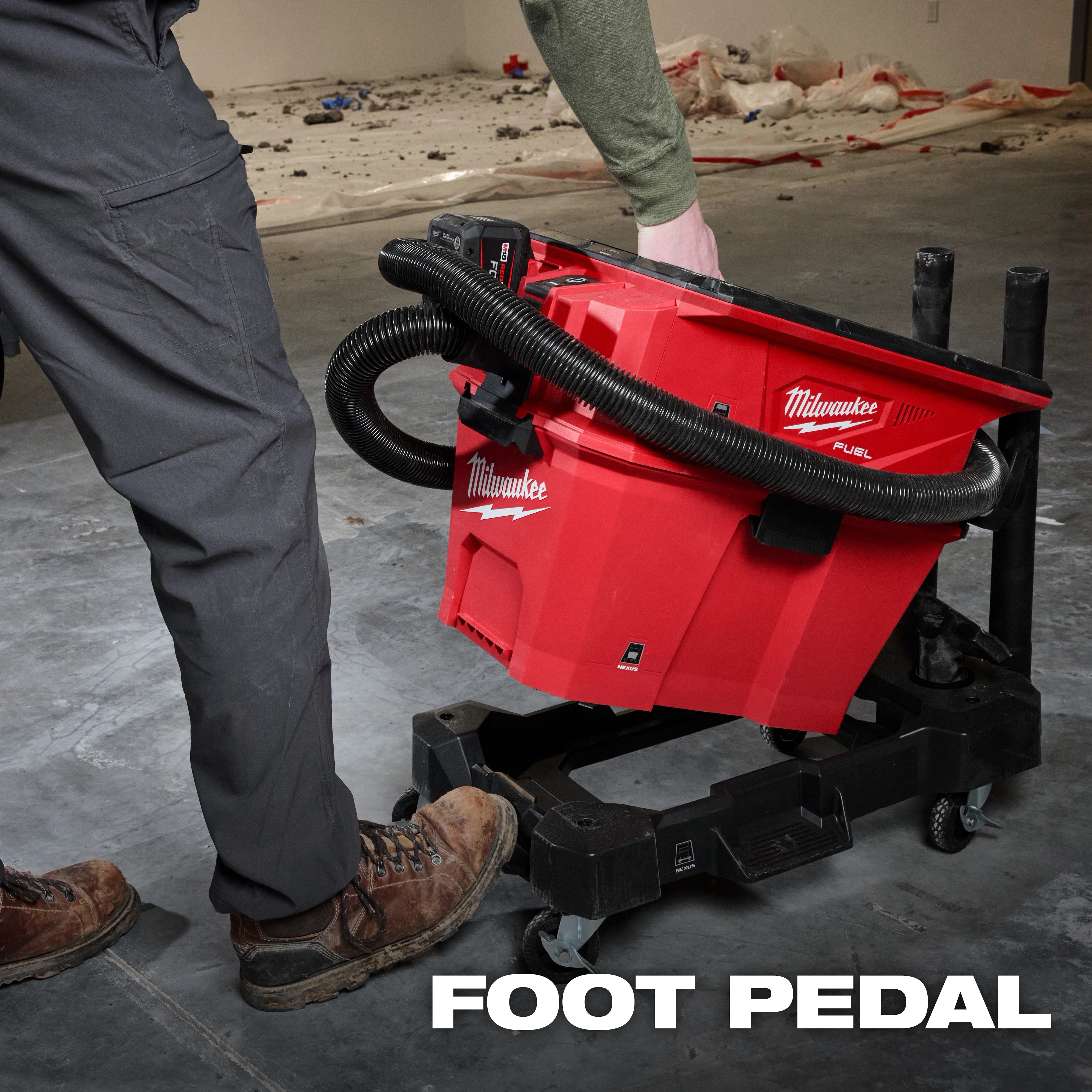 A person operates the NEXUS™ Wet/Dry Vacuum Cart, a red Milwaukee-branded vacuum with a foot pedal and hose attachment, positioned on a wheeled cart in a work area with tools and debris. The text "FOOT PEDAL" is displayed at the bottom.