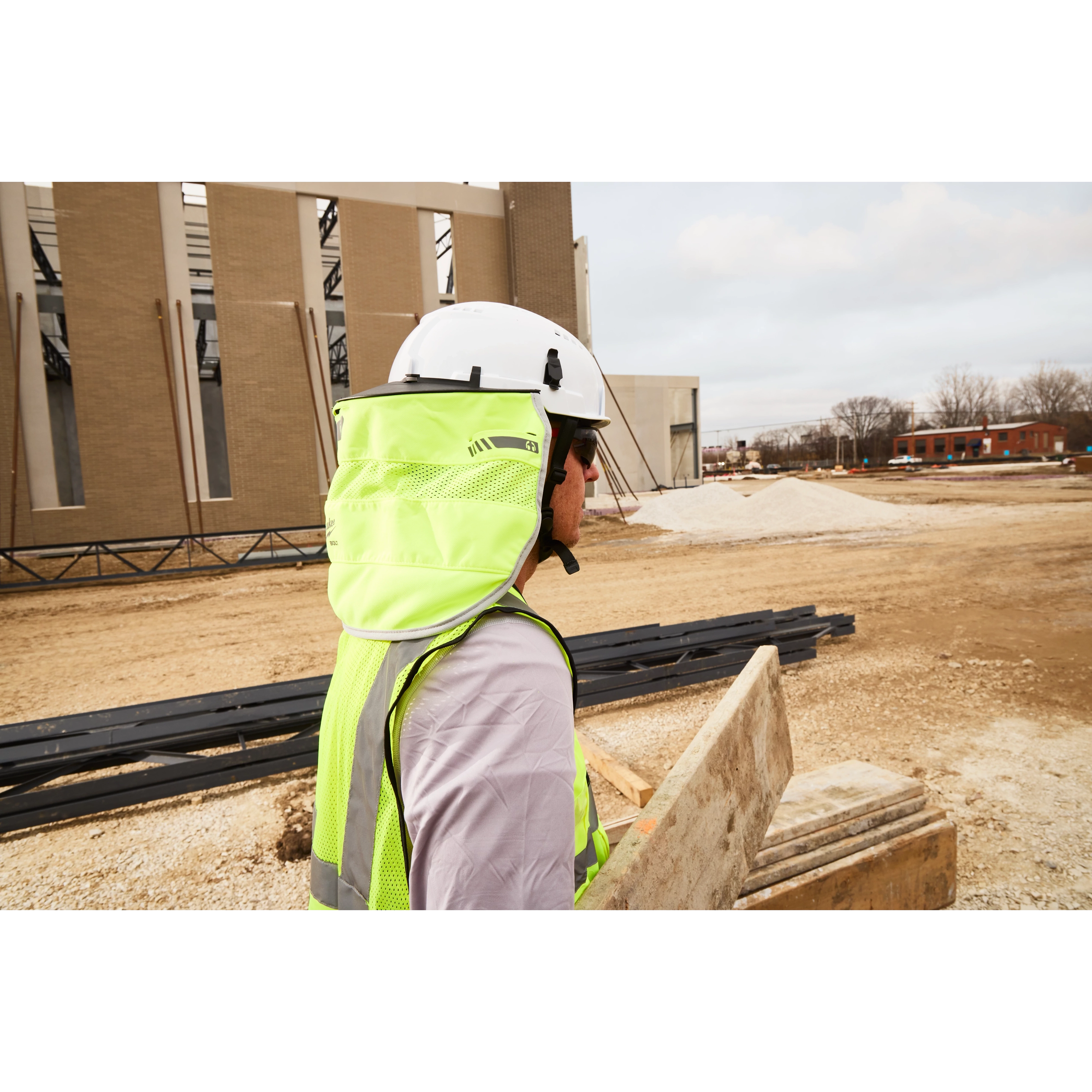 A construction worker wearing a white hard hat, sunglasses, and a neon yellow safety vest with a neck cover stands at a building site looking at structure beams. The ground is covered with dirt and gravel, and an unfinished building is visible in the background.