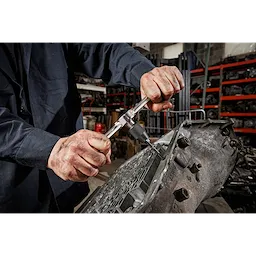 A person uses the Hex-LOK™ 2-in-1 Tap and Die Threading Handle & Tap Collet to work on a metal part in an industrial setting, surrounded by machinery and tools.