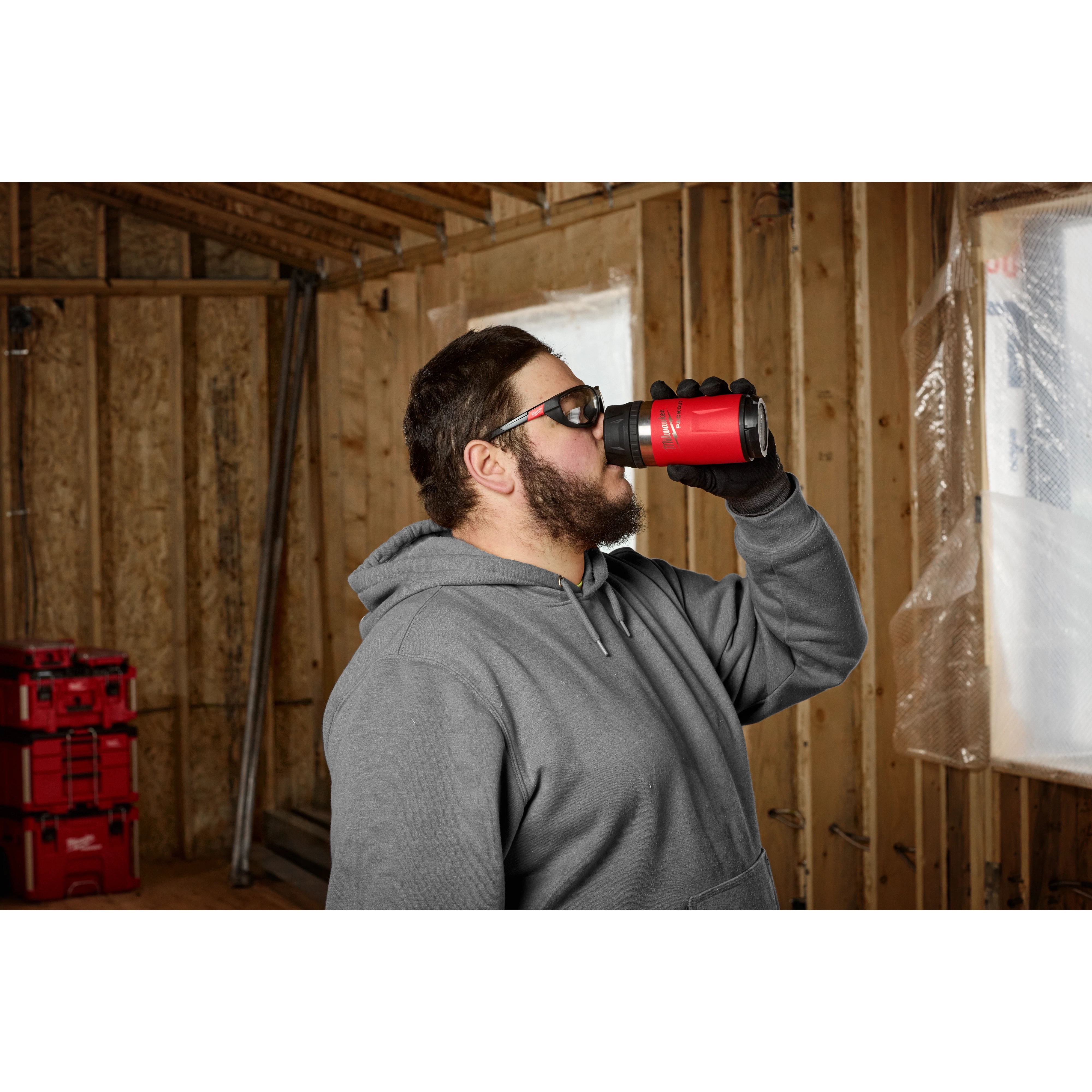 Person drinking from a PACKOUT 12oz Insulated Bottle with Sip Lid in a construction environment. Red tool storage boxes in the background.
