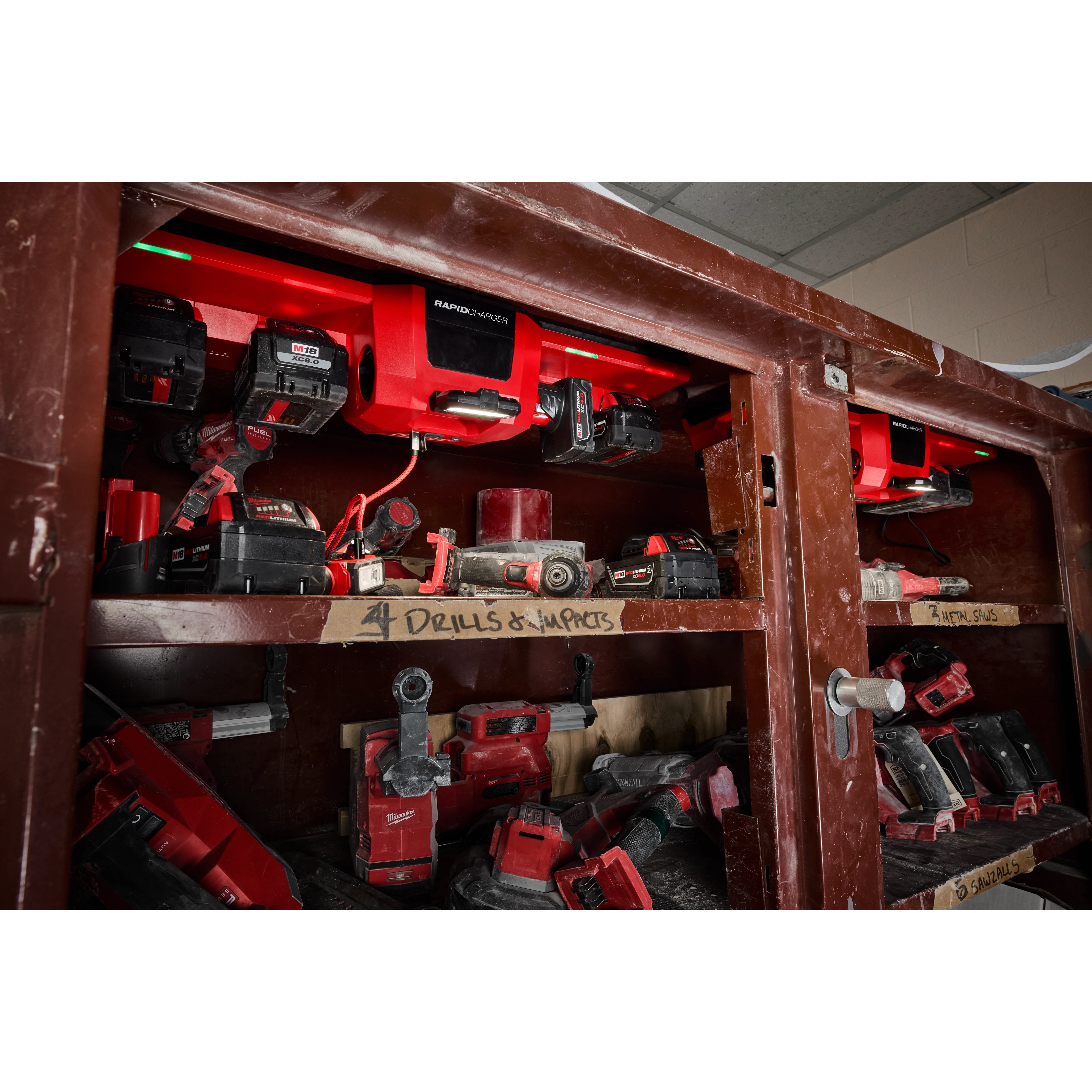 The image displays a storage cabinet containing red and black tools, with the M18™ and M12™ Gangbox Rapid Charger mounted inside the top shelf, charging multiple tool batteries. The shelves are labeled for organized storage of drills, impacts, and saws.