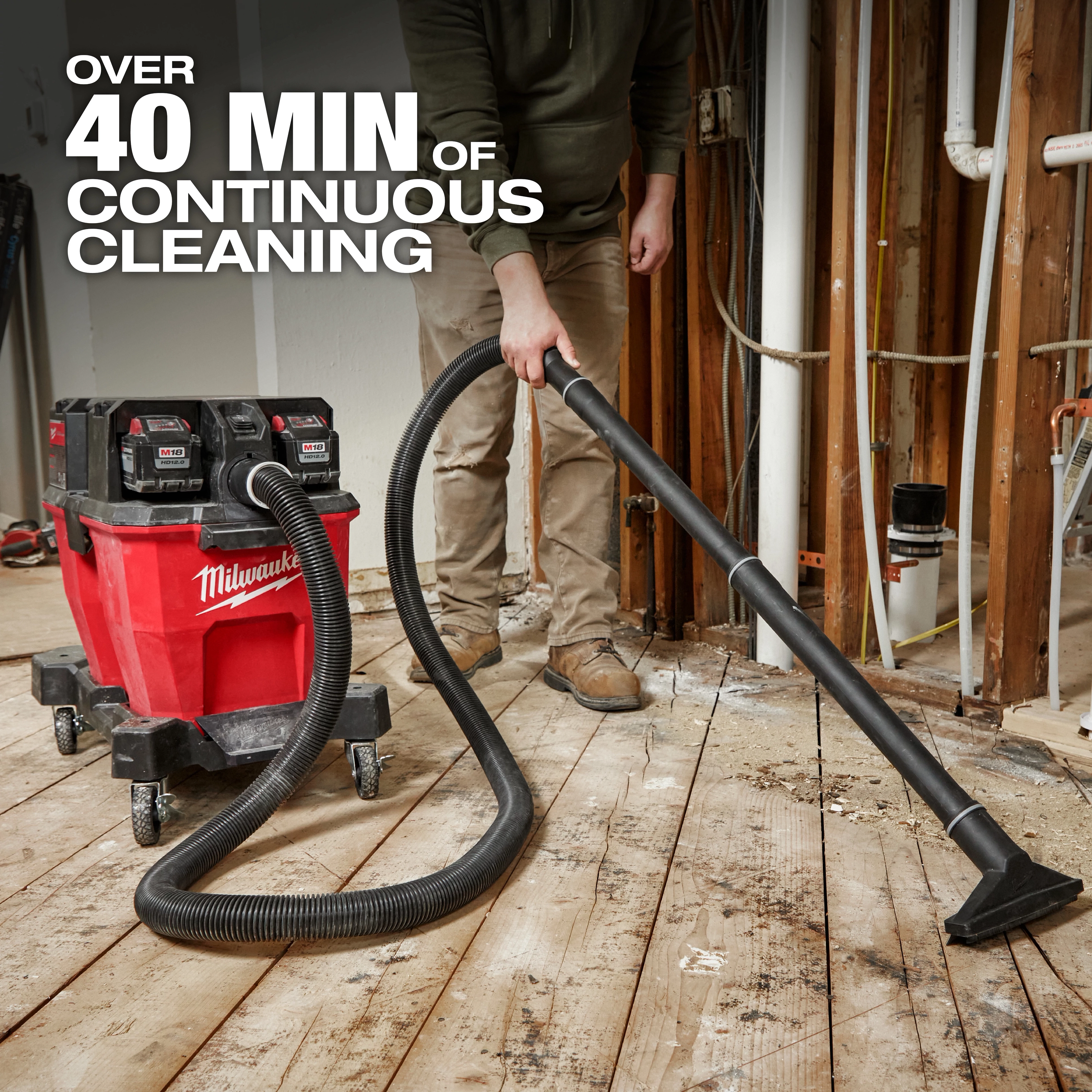 A person uses the M18 FUEL™ NEXUS™ 9 Gallon Dual-Battery Wet/Dry Vacuum, which offers over 40 minutes of continuous cleaning. The red vacuum with black batteries is on wheels and has a long hose being used to clean a wooden floor in a construction area.