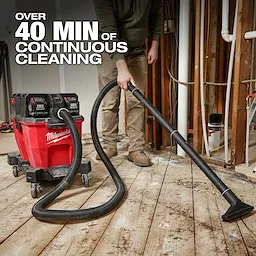 A person uses the M18 FUEL™ NEXUS™ 9 Gallon Dual-Battery Wet/Dry Vacuum, which offers over 40 minutes of continuous cleaning. The red vacuum with black batteries is on wheels and has a long hose being used to clean a wooden floor in a construction area.