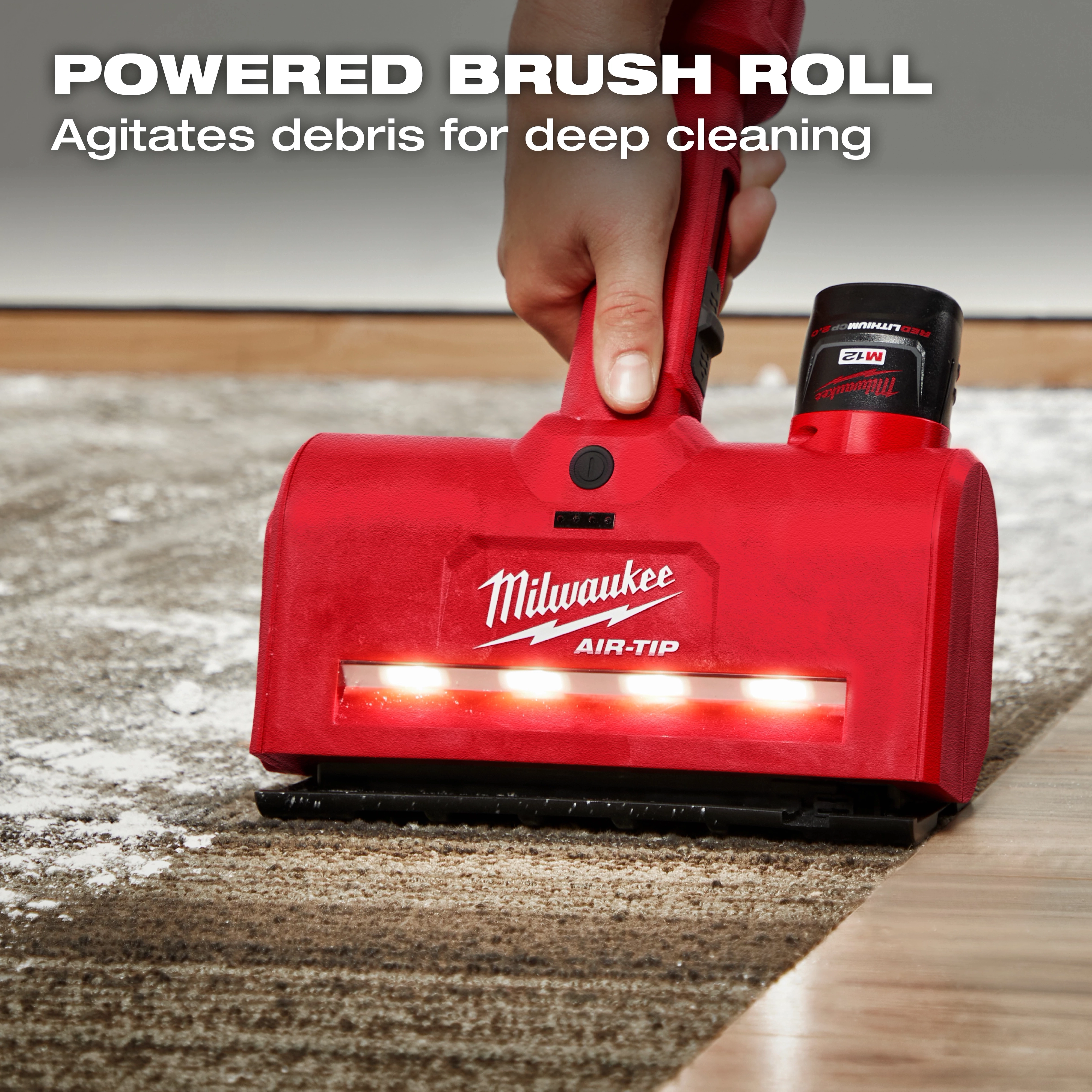 A person uses an M12™ AIR-TIP™ Utility Nozzle, a red handheld carpet cleaning tool by Milwaukee. The nozzle has a powered brush roll and lights, agitating debris for deep cleaning. The text "POWERED BRUSH ROLL Agitates debris for deep cleaning" is displayed at the top.
