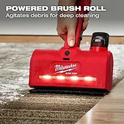 A person uses an M12™ AIR-TIP™ Utility Nozzle, a red handheld carpet cleaning tool by Milwaukee. The nozzle has a powered brush roll and lights, agitating debris for deep cleaning. The text "POWERED BRUSH ROLL Agitates debris for deep cleaning" is displayed at the top.