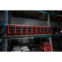 The image shows multiple M18™ Six Bay Daisy Chain Rapid Chargers mounted on a rack in a warehouse. They are charging numerous battery packs simultaneously. The chargers are red and black, and the shelf below holds various tools and equipment.