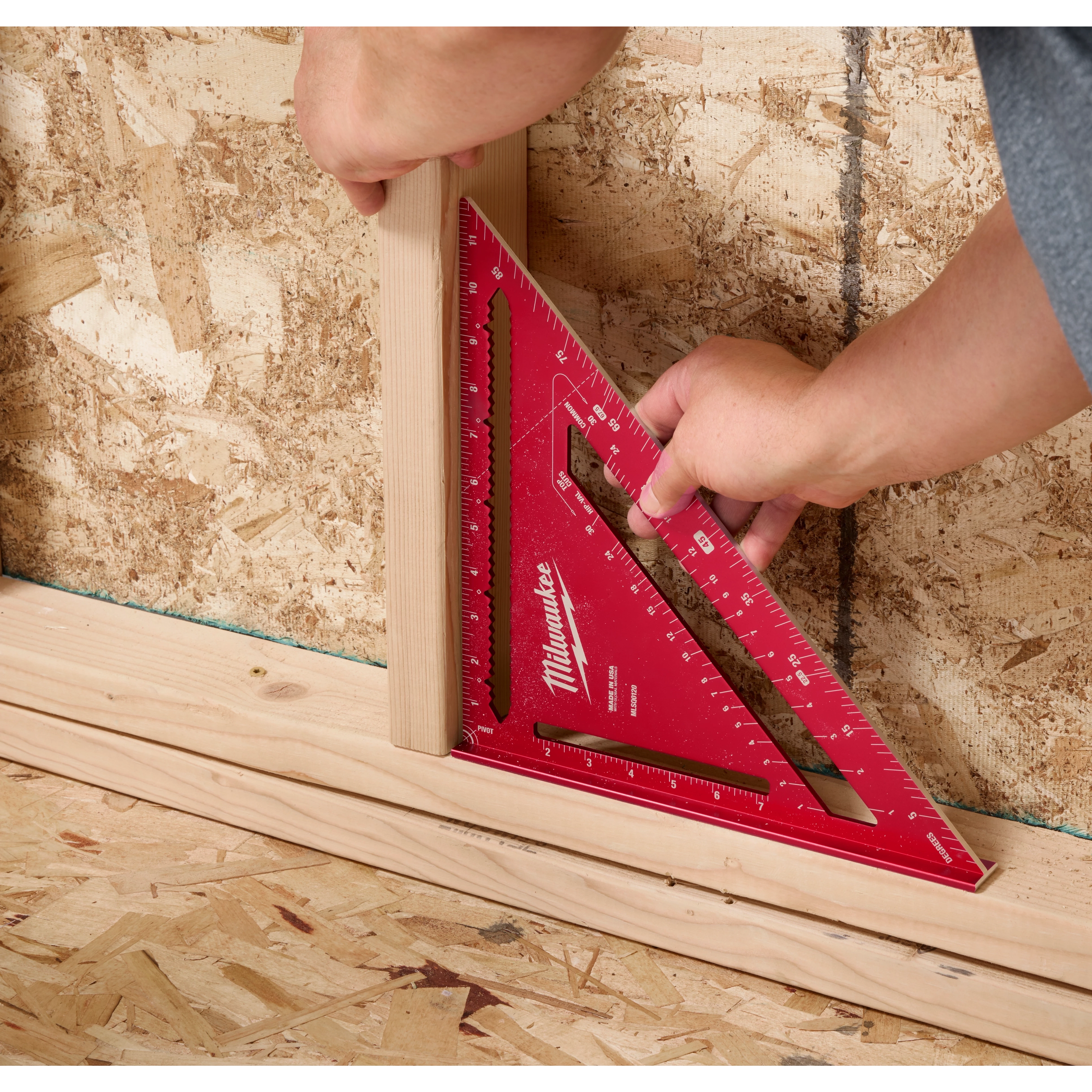 A person uses a red 12" Rafter Square to measure a piece of wood against a plywood wall. The tool features various measurement markings.