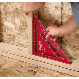A person uses a red 12" Rafter Square to measure a piece of wood against a plywood wall. The tool features various measurement markings.