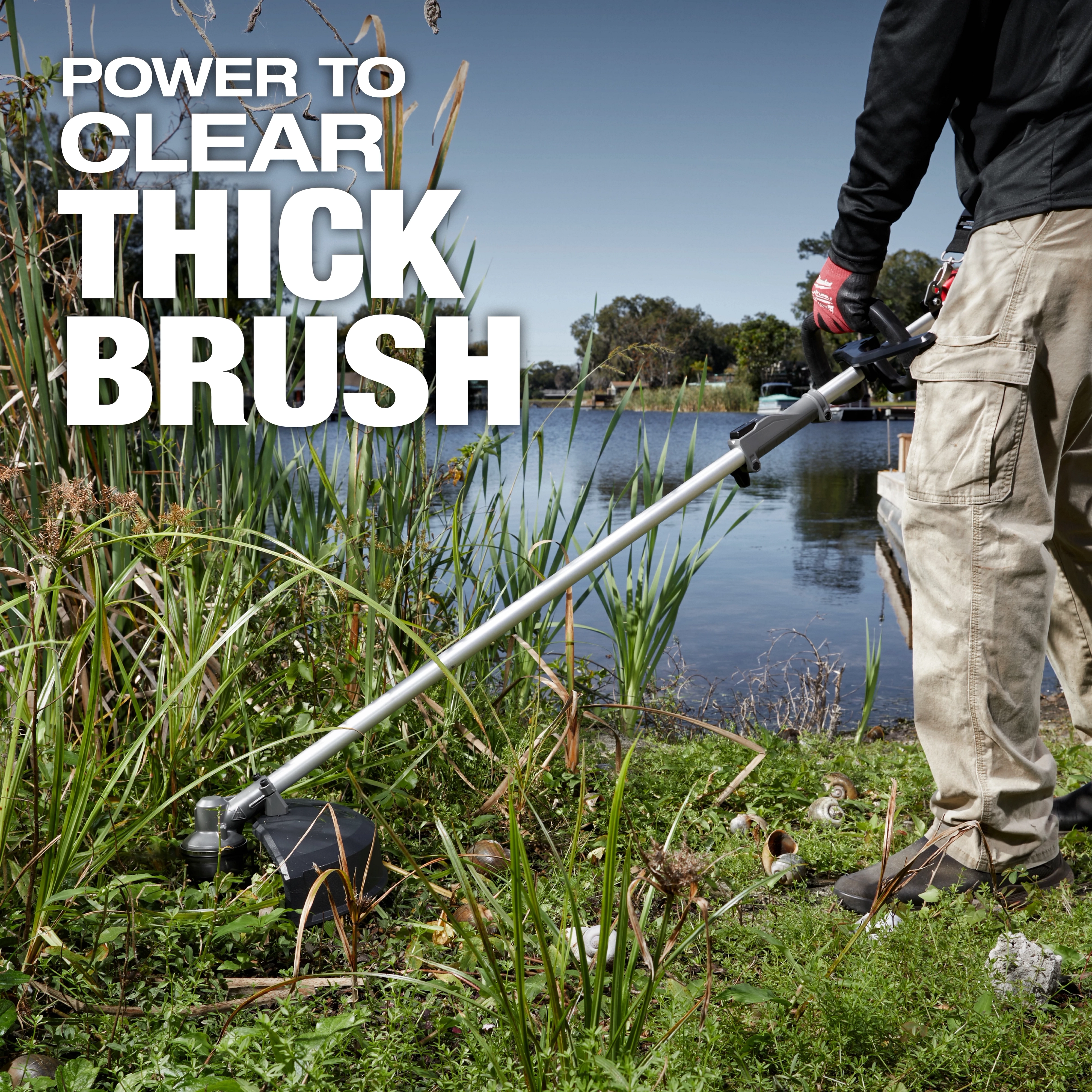 Person using an M18 FUEL QUIK-LOK Brush Cutter Attachment to trim thick brush near a lakeside.
