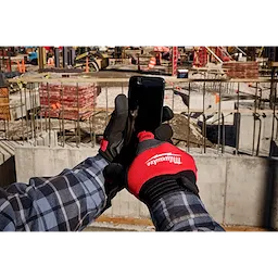 A person wearing a red and black Anti-Vibration Work Glove is holding a smartphone at a construction site. The gloves have the logo of "Milwaukee" on them. The background features construction materials, equipment, and unfinished structures.