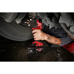 A person is using the M18™ REDLITHIUM™ FORGE™ XC6.0 battery with a power tool to work on a vehicle's wheel. The person is lying on the ground, partially under the vehicle. The battery and tool are highlighted in red and black.