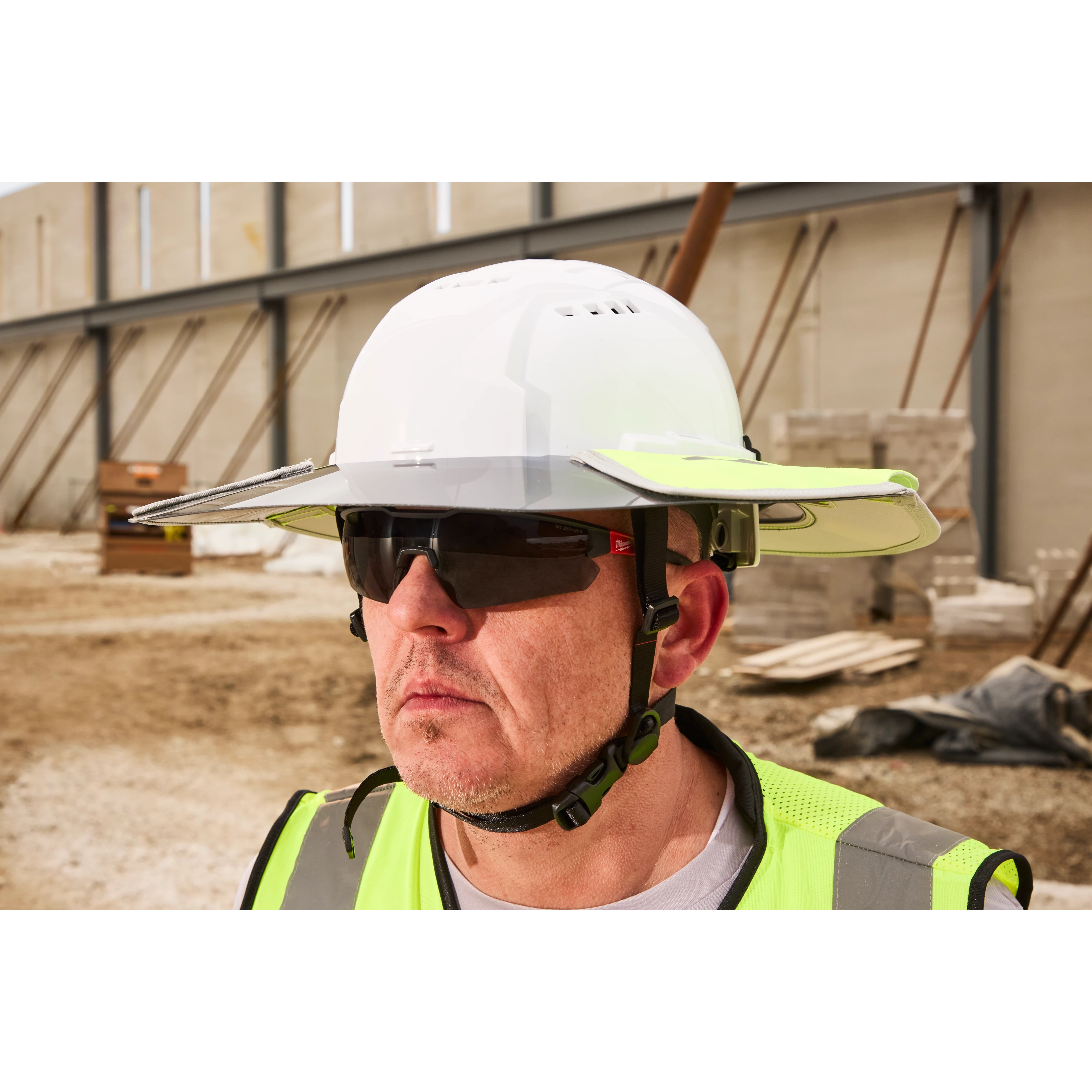 A construction worker wearing a white hard hat and high-visibility yellow safety vest stands in front of an unfinished building. The worker appears to be on a construction site with scattered building materials and structures in the background.