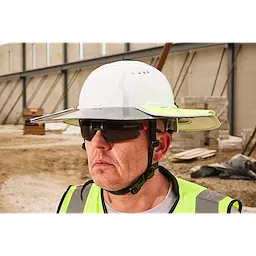 A construction worker wearing a white hard hat and high-visibility yellow safety vest stands in front of an unfinished building. The worker appears to be on a construction site with scattered building materials and structures in the background.