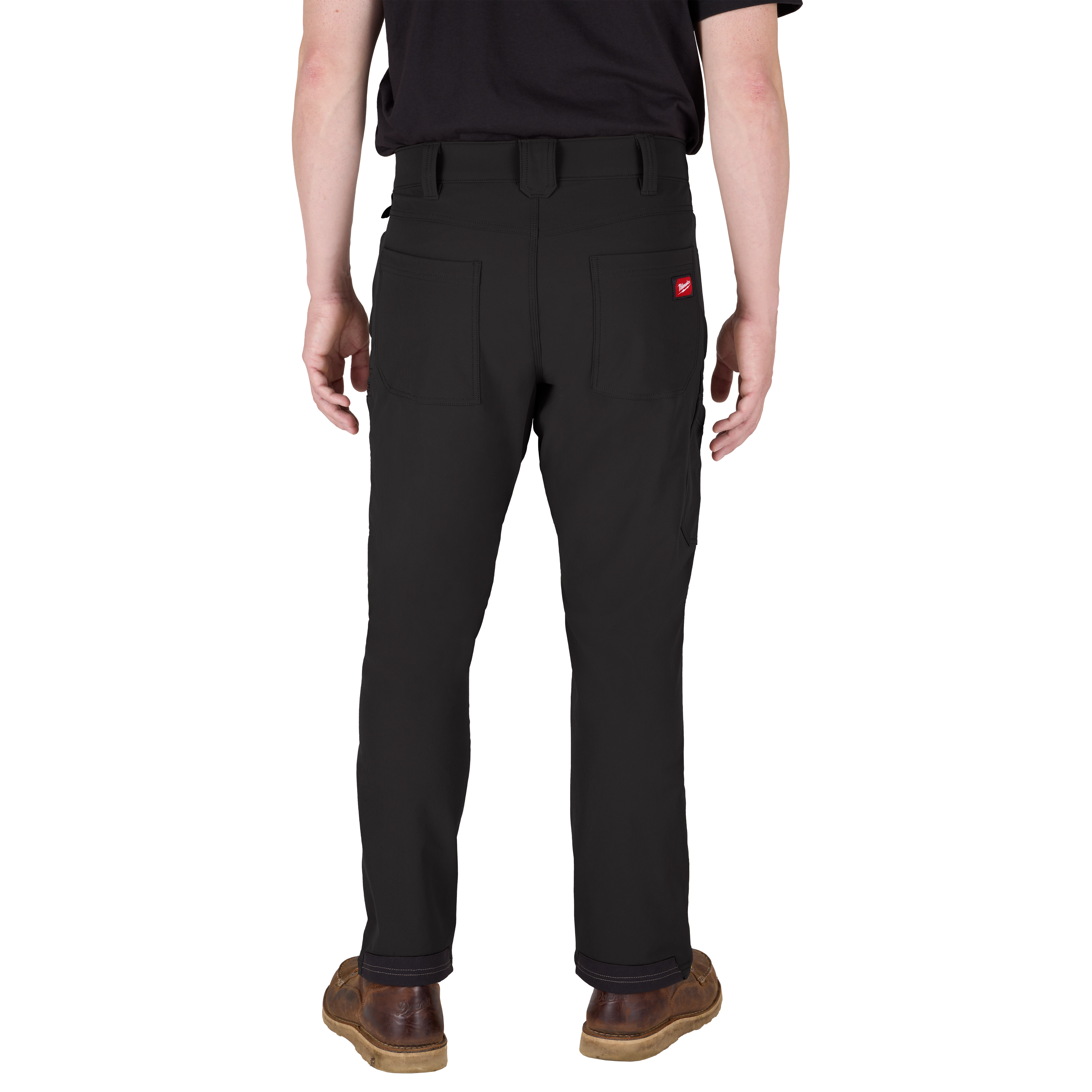 A person is standing and facing away from the camera, wearing black pants with rear pockets and a black shirt. The person has a small, red logo tag on the right rear pocket of the pants. They are also wearing brown shoes.