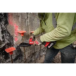 A person wearing a neon green jacket and gloves is using an M18 FUEL™ #10 (1-1/4") Rebar Cutter to cut through rebar protruding from a concrete wall. The rebar segments are topped with orange plastic caps for safety.