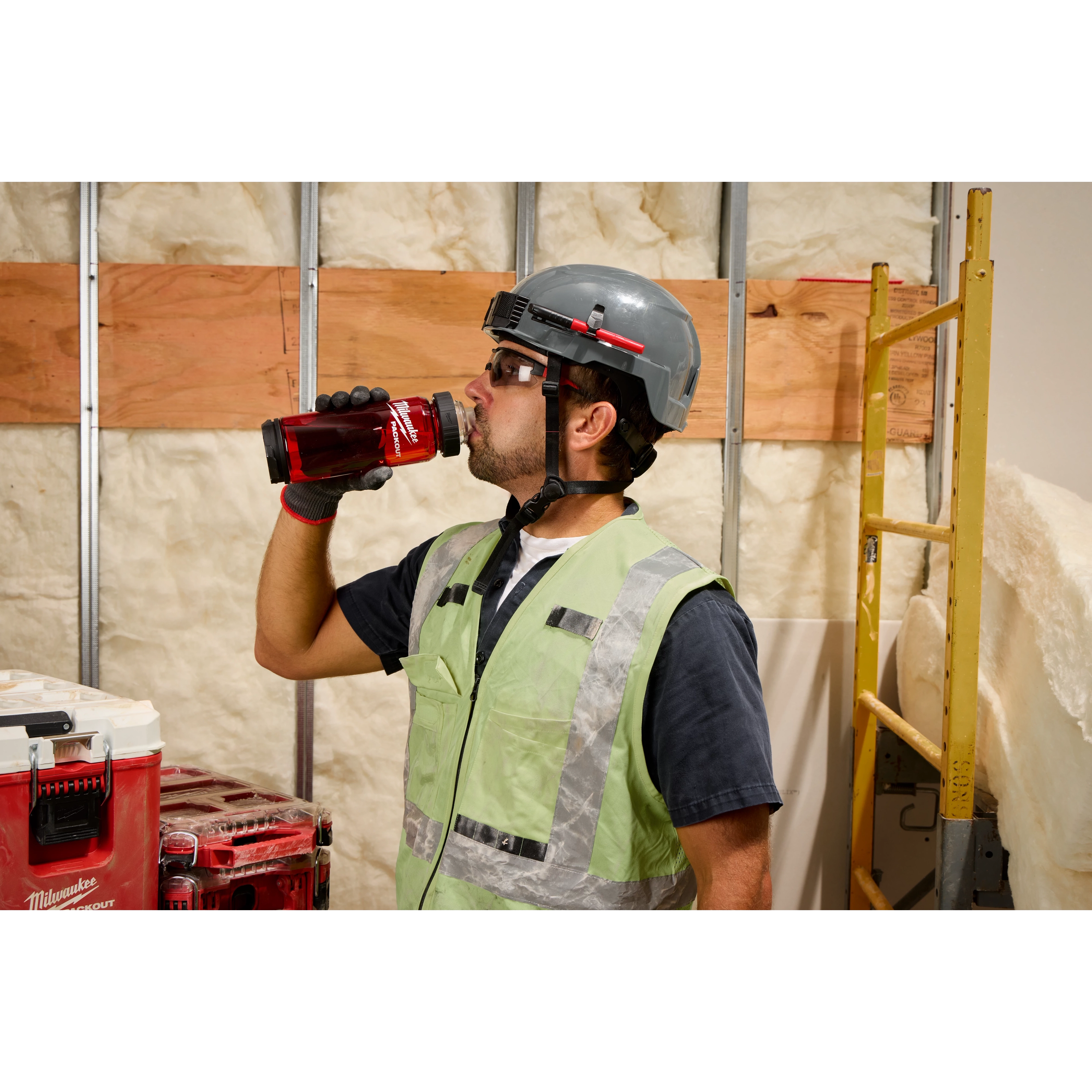 A construction worker wearing a gray helmet and reflective vest is drinking from a red PACKOUT™ 25oz Bottle with Chug Lid. The background shows an unfinished wall with insulation, wooden planks, and a yellow ladder. A stack of PACKOUT™ tool cases is visible to the left.