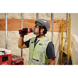 A construction worker wearing a gray helmet and reflective vest is drinking from a red PACKOUT™ 25oz Bottle with Chug Lid. The background shows an unfinished wall with insulation, wooden planks, and a yellow ladder. A stack of PACKOUT™ tool cases is visible to the left.