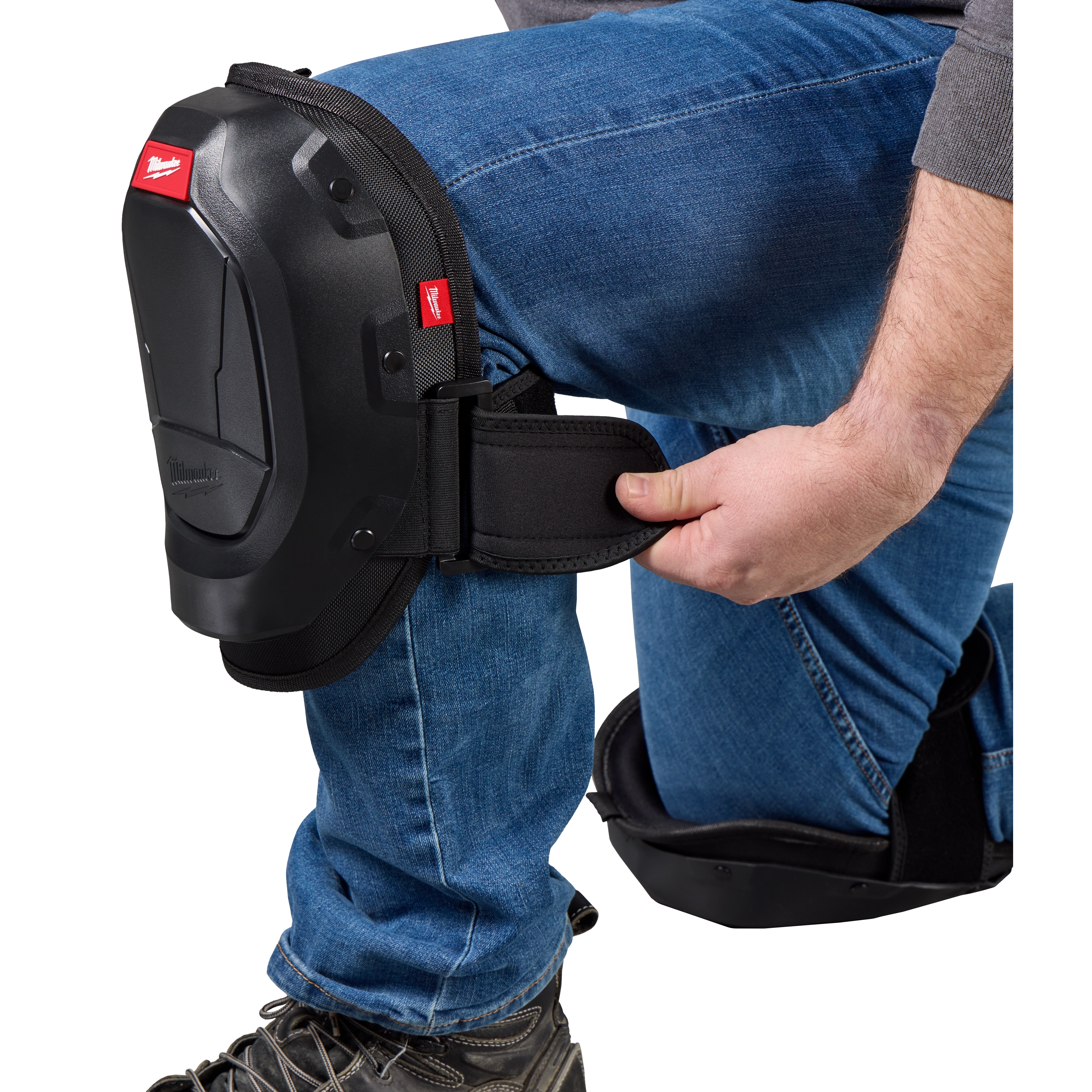A person wearing jeans is adjusting a black knee pad on their bent knee. The knee pad has a small Milwaukee Tool logo on it. The person is holding the strap of the knee pad, ensuring it is secure. The image highlights the knee pad's protective design.