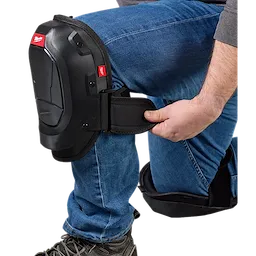 A person wearing jeans is adjusting a black knee pad on their bent knee. The knee pad has a small Milwaukee Tool logo on it. The person is holding the strap of the knee pad, ensuring it is secure. The image highlights the knee pad's protective design.
