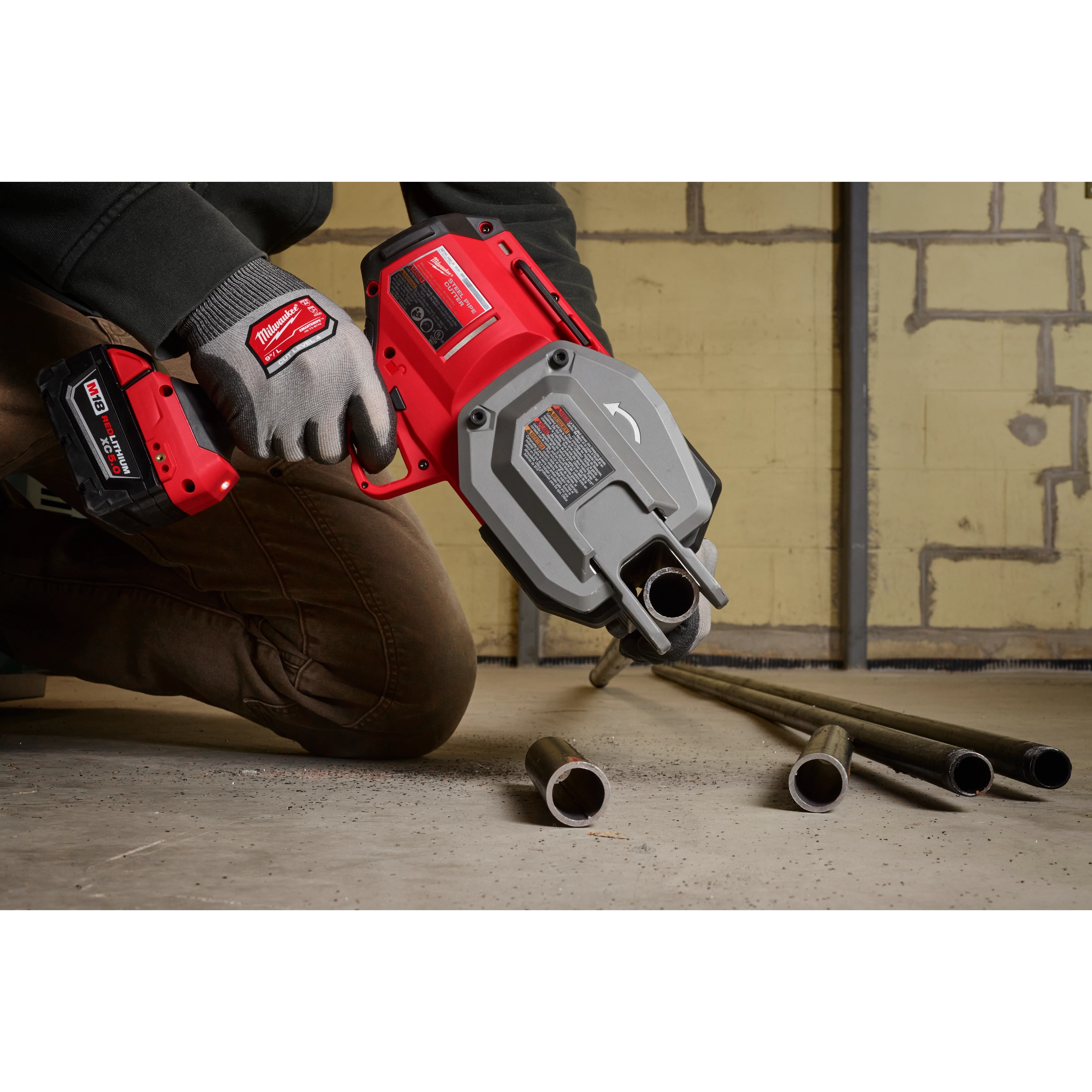 A person is using the M18 FUEL™ ½”-1” Steel Pipe Cutter to cut metal pipes on a concrete floor. The tool is red and gray, and the user is wearing gloves with a matching branding logo.