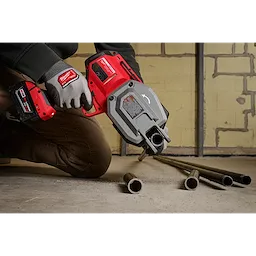 A person is using the M18 FUEL™ ½”-1” Steel Pipe Cutter to cut metal pipes on a concrete floor. The tool is red and gray, and the user is wearing gloves with a matching branding logo.