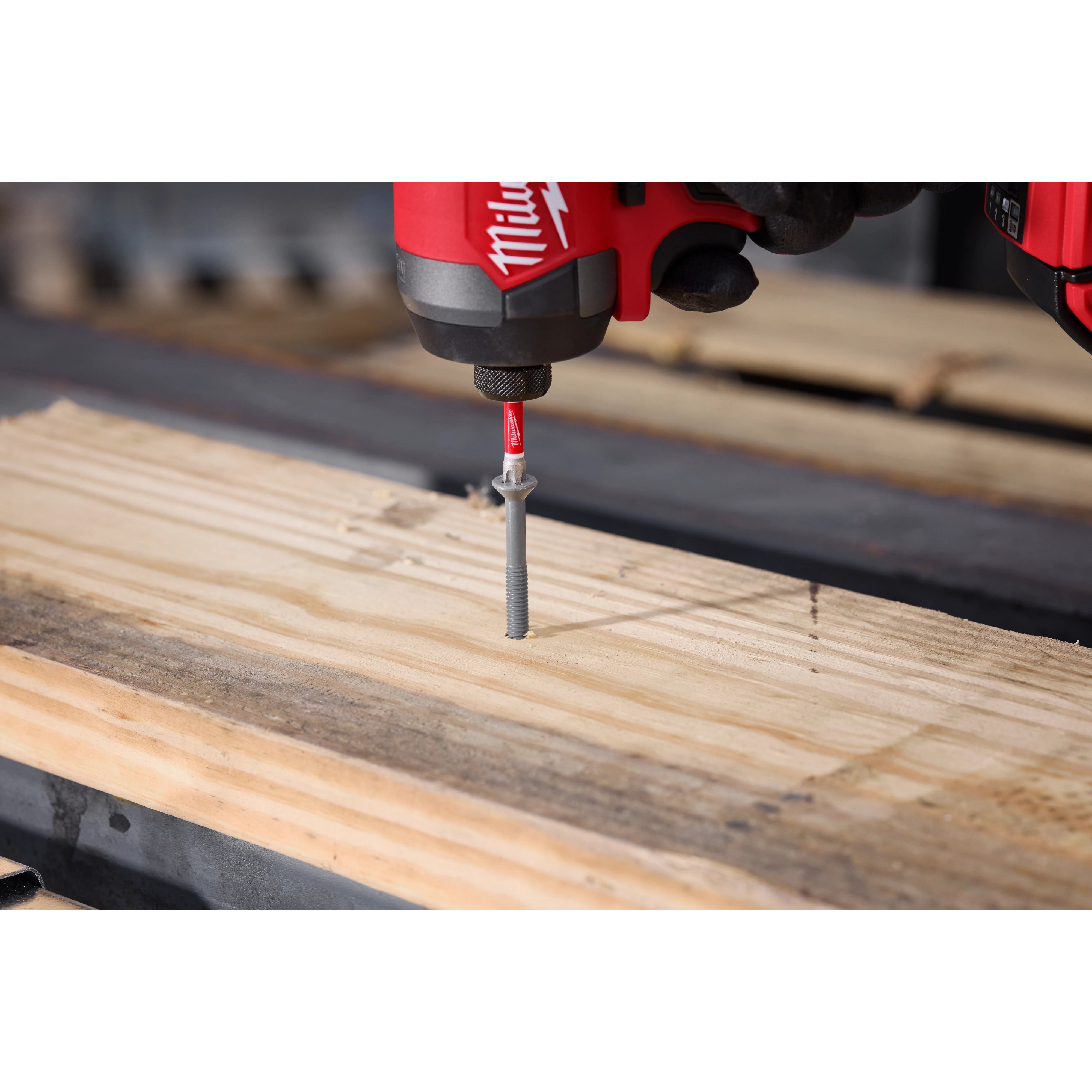 A close-up of a red power drill driving a screw into a piece of wood using the SHOCKWAVE™ 2” Impact Phillips #3 Power Bit. The drill is shown in action, firmly holding and maneuvering the screw into the plank.