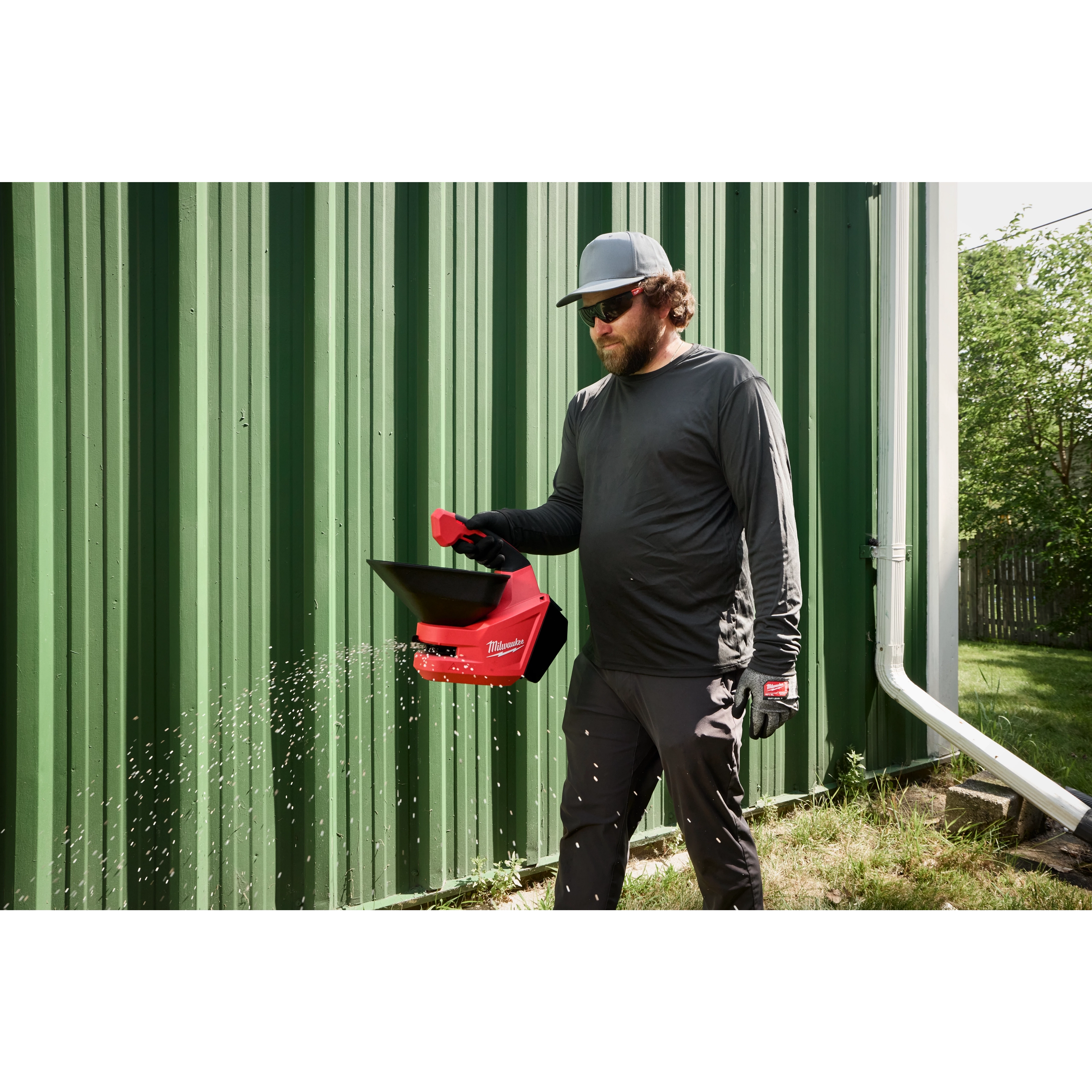 Person walking on grass using a red M12 Handheld Broadcast Spreader beside a green metal building.