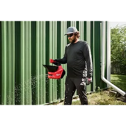 Person walking on grass using a red M12 Handheld Broadcast Spreader beside a green metal building.
