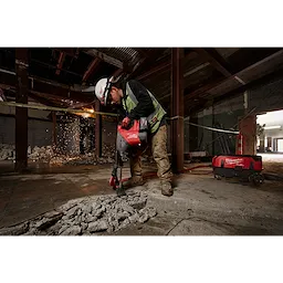 A construction worker in safety gear uses the M18 FUEL(tm) 1-3/4" SDS MAX Rotary Hammer Kit w/ONE-KEY to break concrete in a partially demolished building. Sparking debris and a red toolbox are visible in the background.