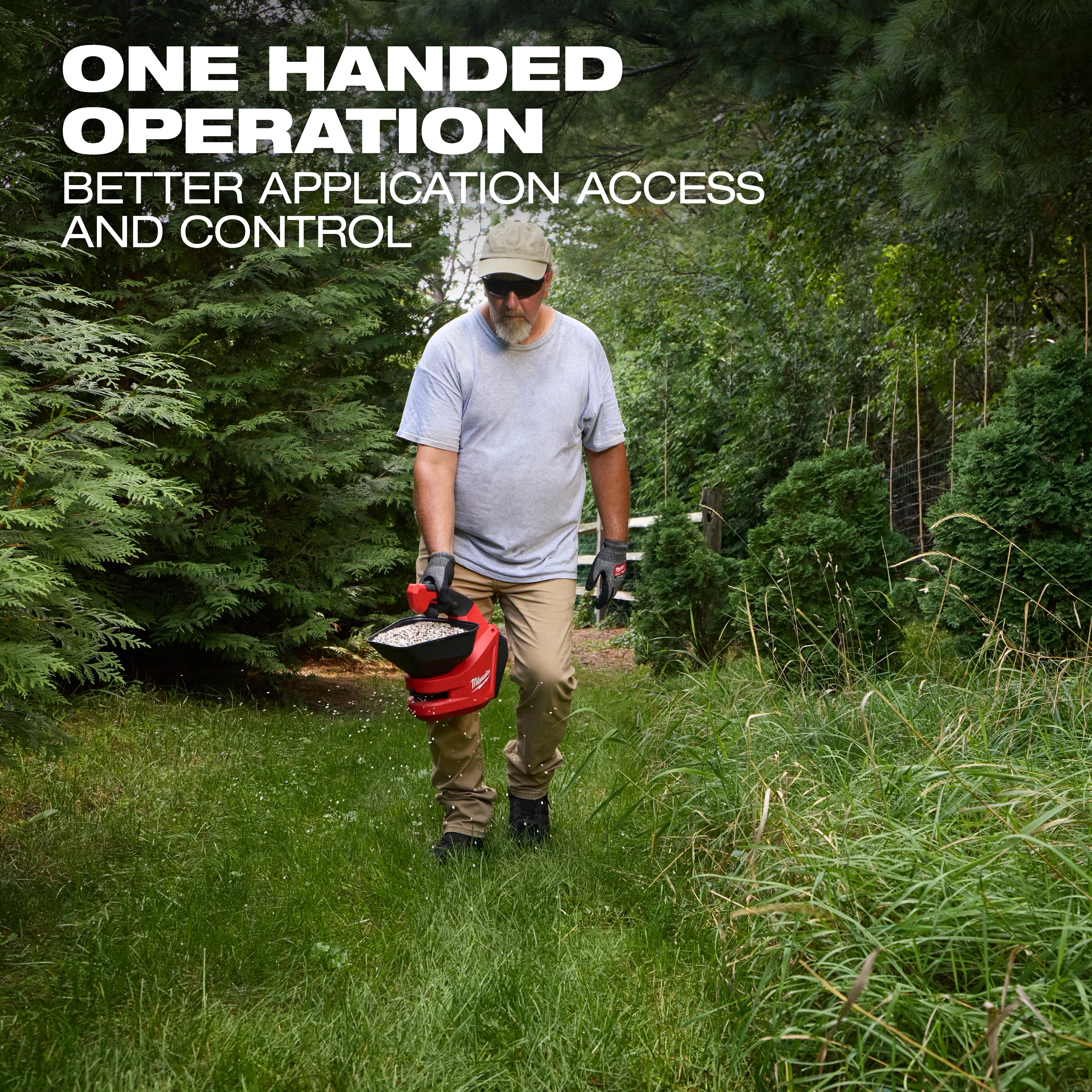 Person walking on grass holding an M12 Handheld Broadcast Spreader in one hand among tall trees and bushes.