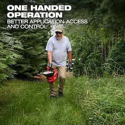 Person walking on grass holding an M12 Handheld Broadcast Spreader in one hand among tall trees and bushes.