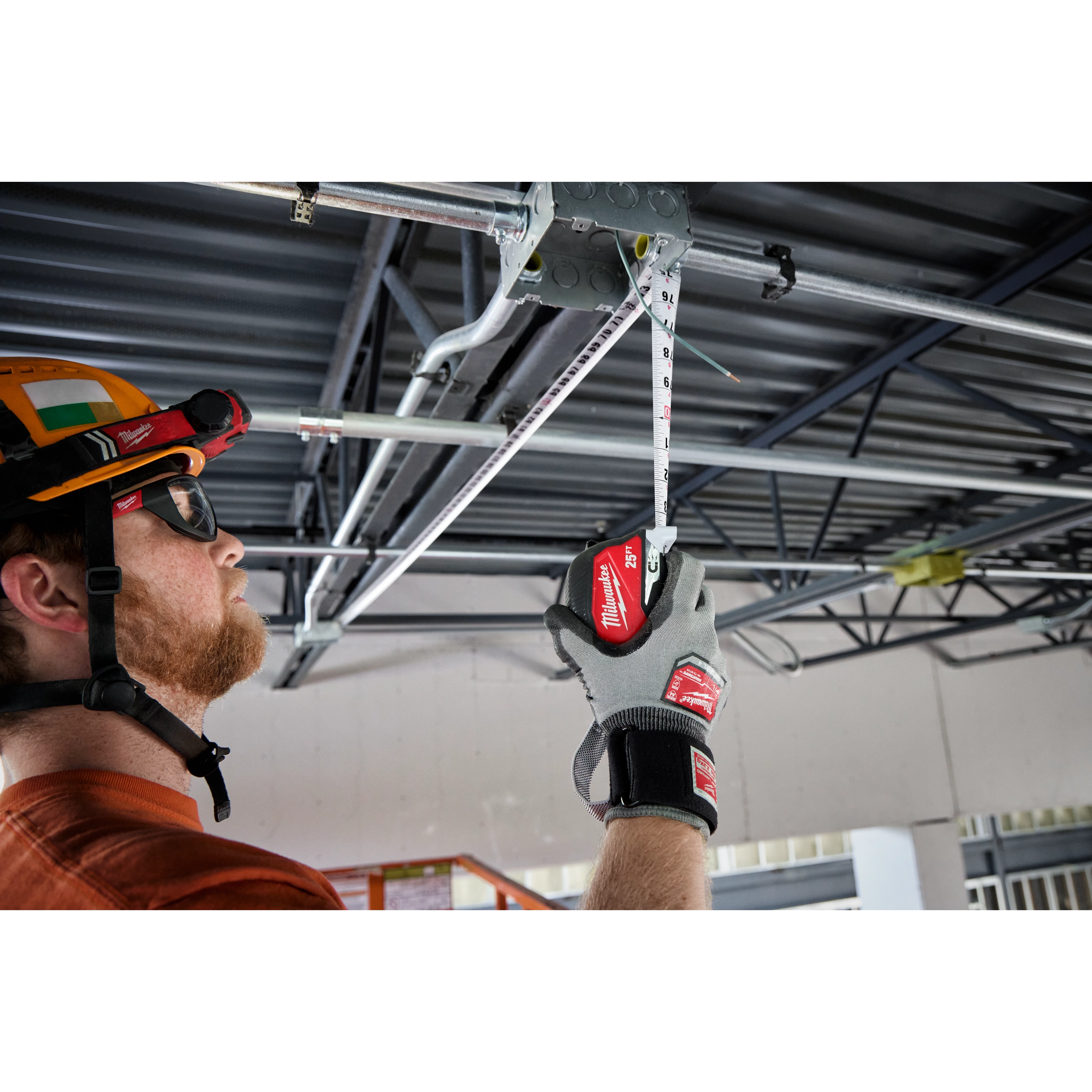 A person wearing gloves is using a 25ft Electrician's Magnetic Tape Measure to measure piping attached to a metal beam on a ceiling. The tape measure is extended and held in place, and the person is positioned on the left side of the image.