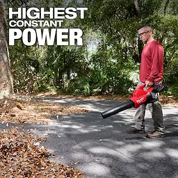 A person is using an M18 FUEL™ Dual Battery Blower to clear leaves from a paved path surrounded by trees. The text "HIGHEST CONSTANT POWER" is displayed at the top left of the image.