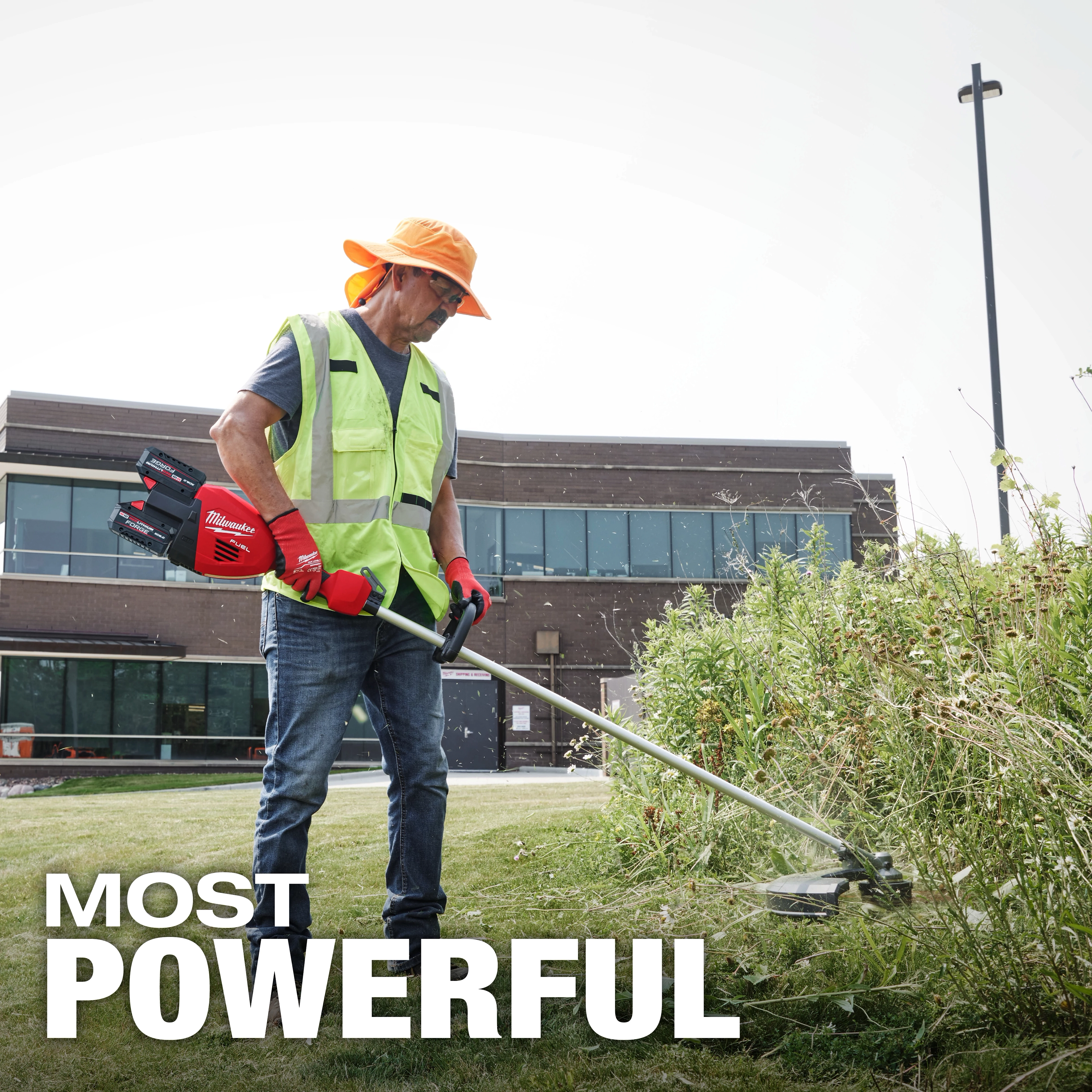 A person wearing a high-visibility vest, orange hat, and red gloves is using an M18 FUEL 17” Dual Battery String Trimmer to trim overgrown grass near a building. The text "MOST POWERFUL" is displayed at the bottom of the image.