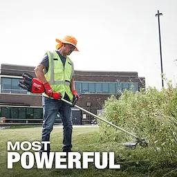 A person wearing a high-visibility vest, orange hat, and red gloves is using an M18 FUEL 17” Dual Battery String Trimmer to trim overgrown grass near a building. The text "MOST POWERFUL" is displayed at the bottom of the image.