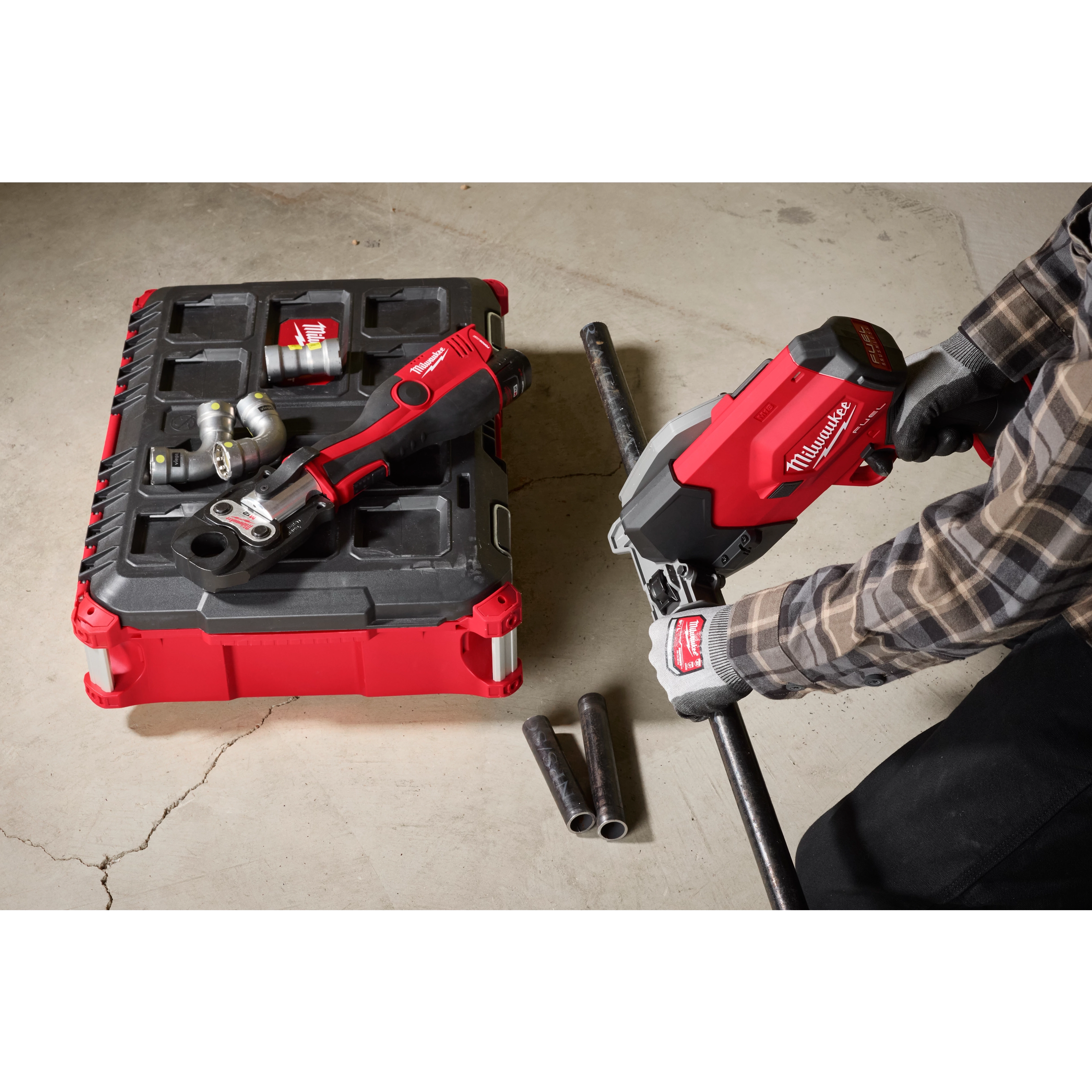 A person uses a Milwaukee M18 FUEL™ ½”-1” Steel Pipe Cutter to cut a pipe on a concrete floor. The tool is red and black, with gloves and other equipment visible in the background.