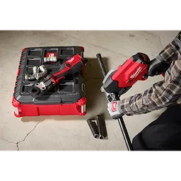 A person uses a Milwaukee M18 FUEL™ ½”-1” Steel Pipe Cutter to cut a pipe on a concrete floor. The tool is red and black, with gloves and other equipment visible in the background.