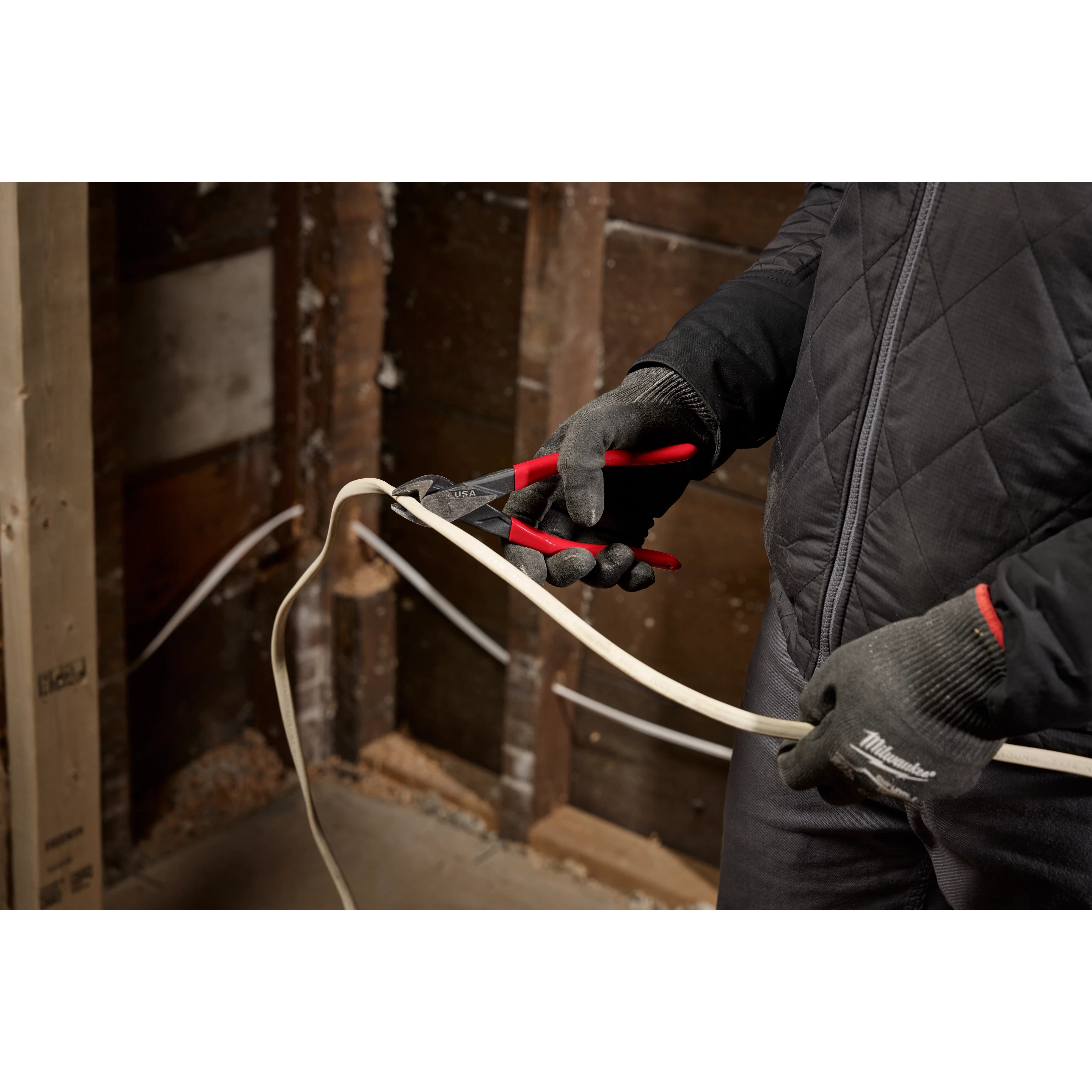 A person wearing gray gloves and a black jacket is using red-handled pliers to cut an electrical wire inside an unfinished wooden wall.