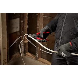 A person wearing gray gloves and a black jacket is using red-handled pliers to cut an electrical wire inside an unfinished wooden wall.