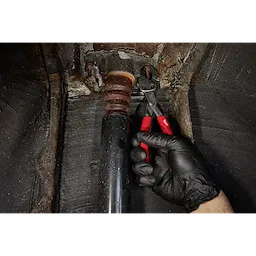 A hand wearing a black glove grips the 8" Slip Joint Pliers to adjust a metal object inside a black pipe. The pliers have a silver head and red handles. The background shows a rusty, textured surface.