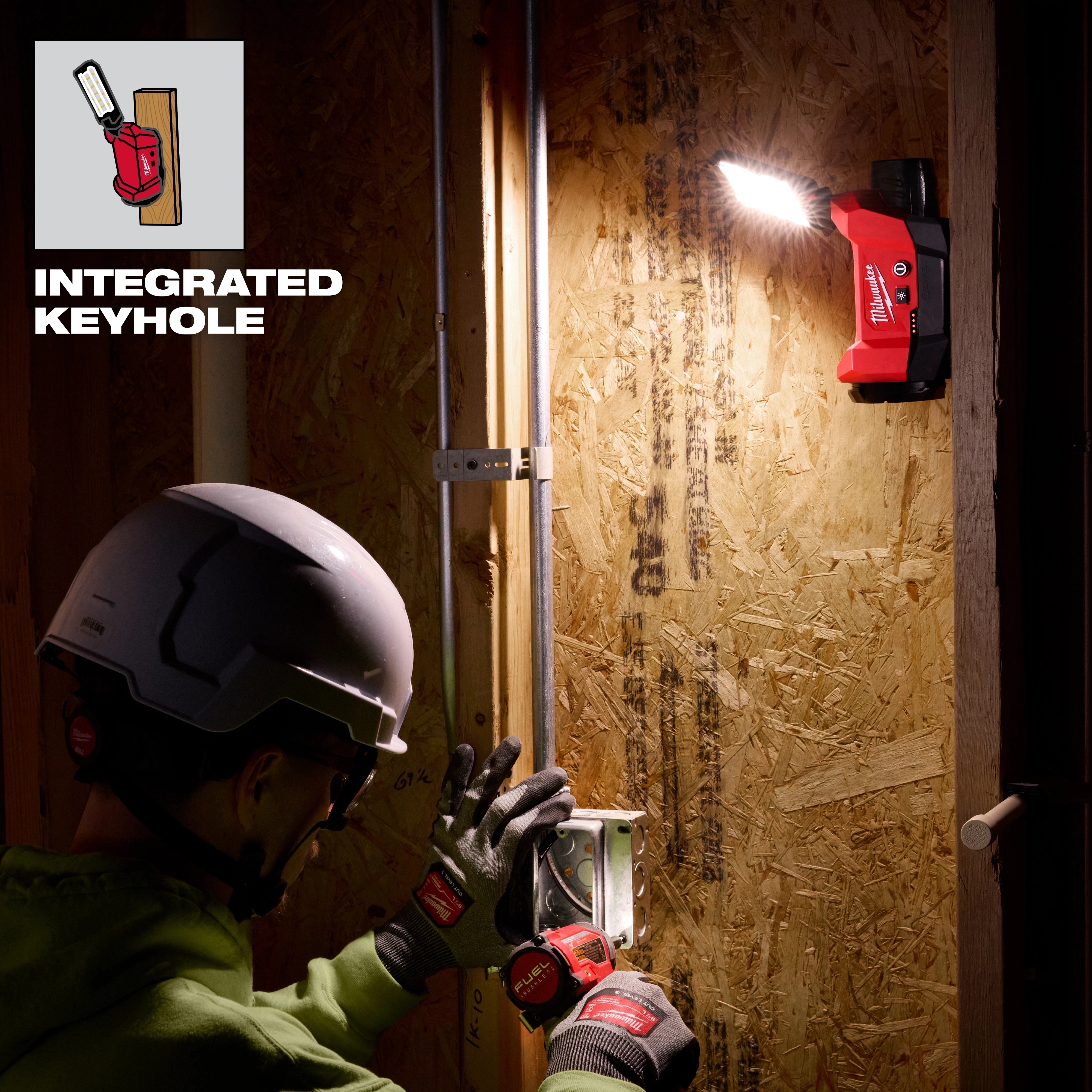 M12™ ROVER™ Multi-Directional Flood Light w/ PACKOUT™ Compatibility illuminates a work area as a person wearing a hard hat and gloves installs an electrical outlet. The flood light is mounted on a wooden panel via an integrated keyhole. The light beam highlights the working space efficiently.